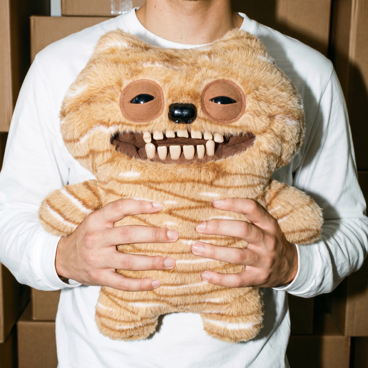 Person holding a Fuggler Bigg Fugg Chuckles 18-Inch Plush featuring fuzzy tan and white striped fur, a wide mouth with human-like teeth, and heavy-lidded eyes.