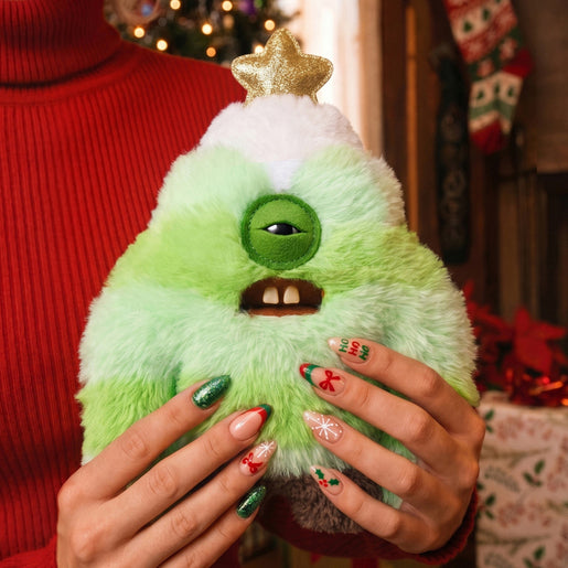 A person in a red sweater holds a fuzzy green monster plush shaped like a Christmas tree, with a gold star on top and holiday nail designs visible.