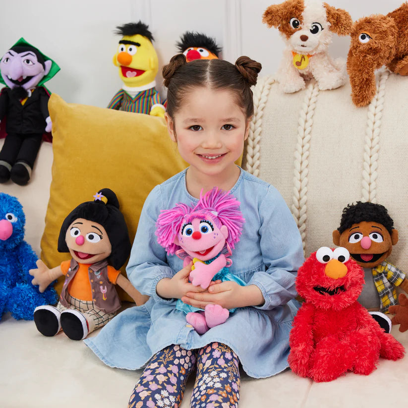 Child holding a pink plush toy surrounded by various Sesame Street characters on a couch.