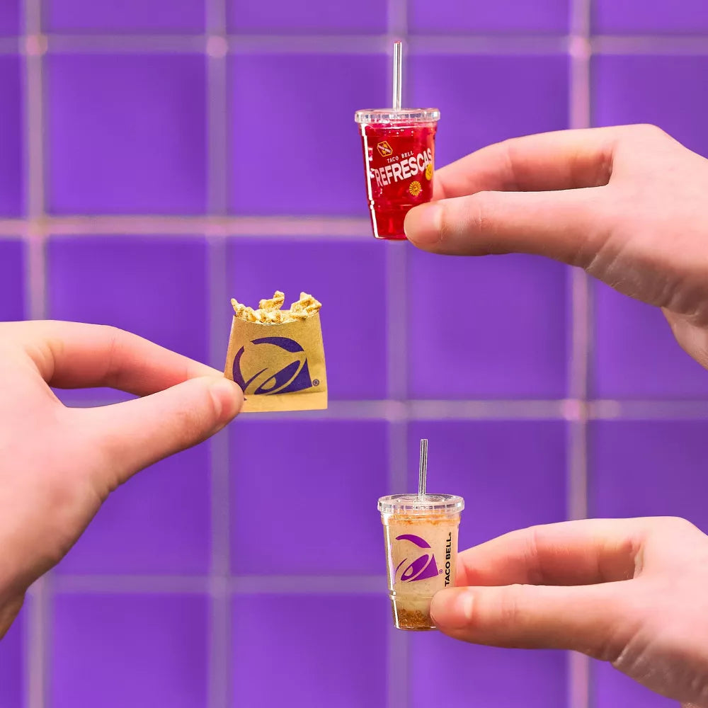 Miniature food items including a drink and a bag held by hands against a purple tiled background.