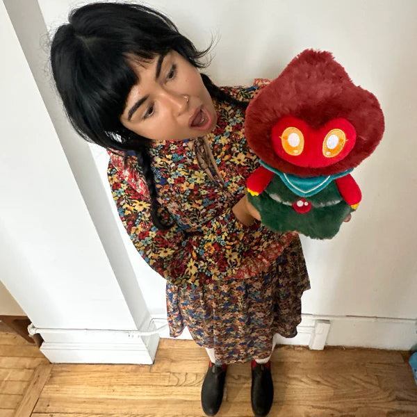 Person holding a Squishable Mini Flatwoods Monster plush toy with a colorful dress indoors.