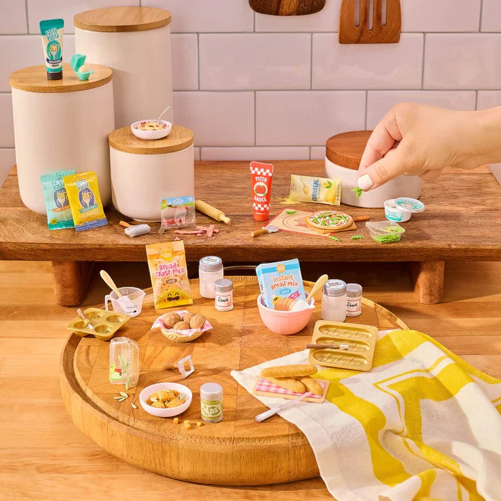 Miniature food items and kitchen accessories on a wooden table with a hand interacting with them. Miniverse Make It Mini From Scratch Series 2