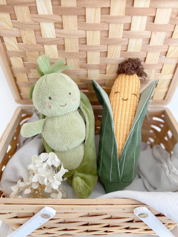 Two plush toys resembling a green vegetable and a corn on a woven basket with white flowers.