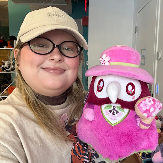 Person wearing a beige cap and glasses holding a plush toy with a pink hat