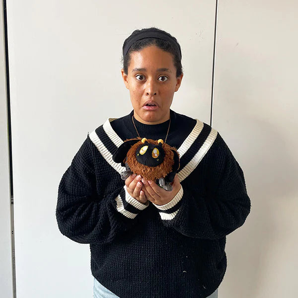 Person wearing a black sweater with white trim holding a Squishable Snackers Death's-head Hawkmoth plush against a plain background
