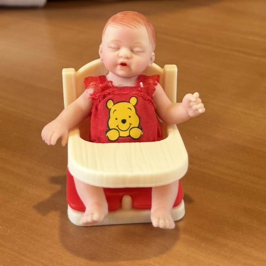 Toy baby figure in a high chair with a red outfit featuring a bear design on a wooden surface.