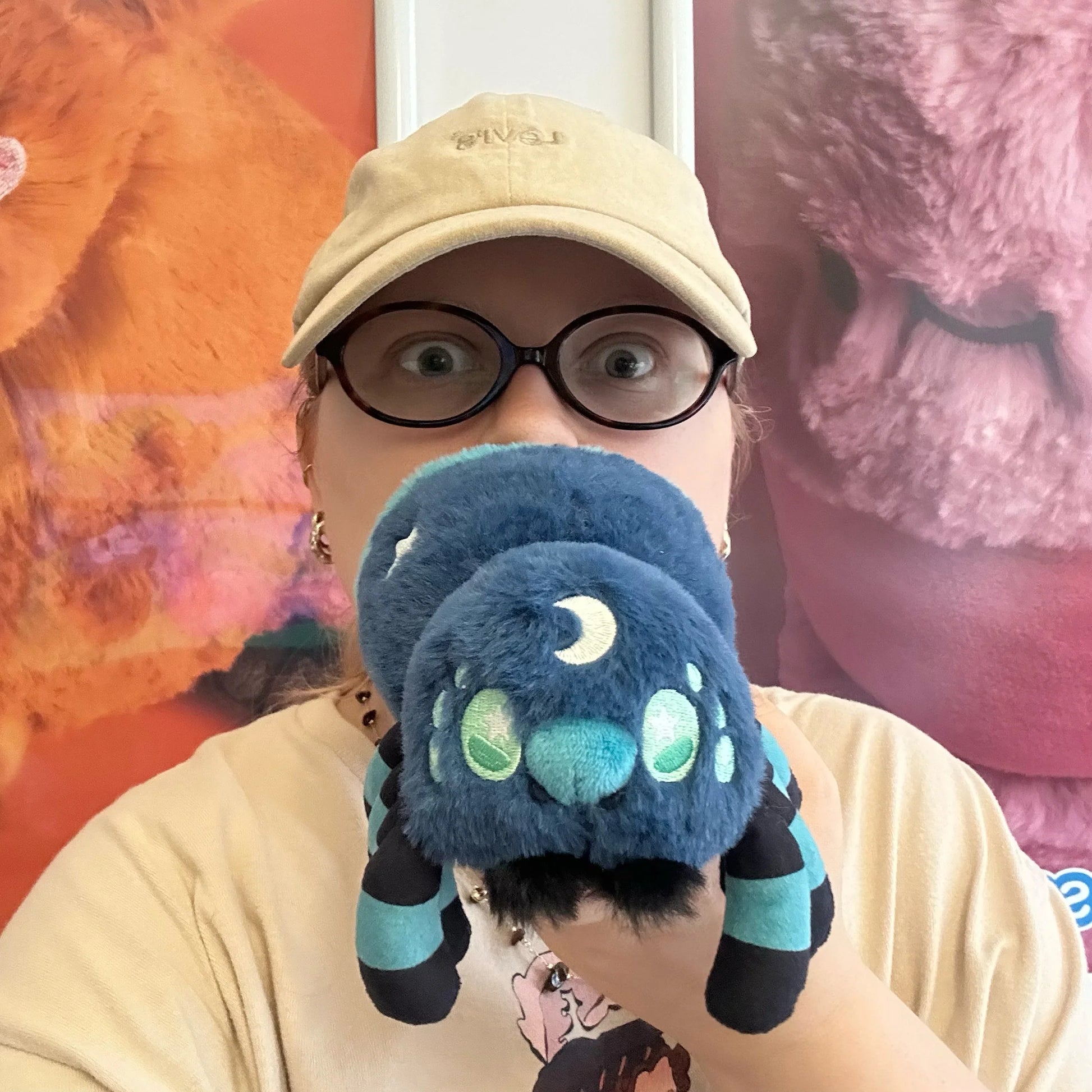 Person wearing a beige cap and glasses, holding a blue plush toy with a colorful background