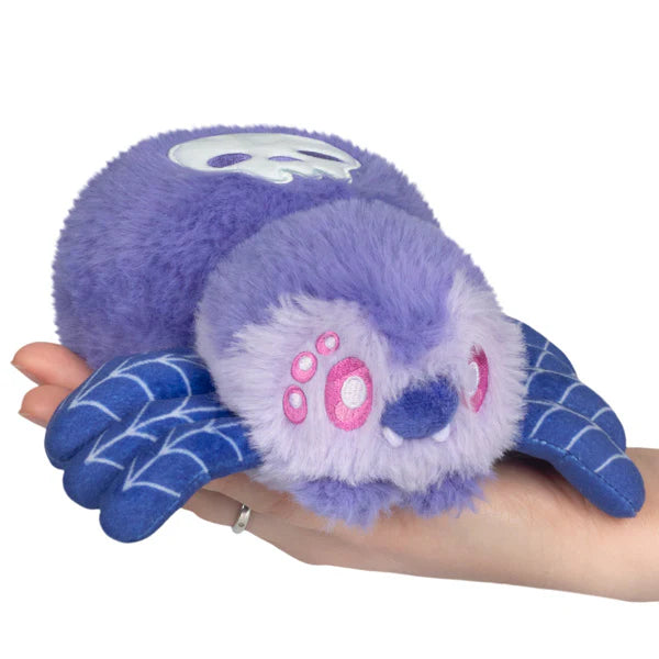 Squishable Alter Egos Series 20 Skull Spider plush held in a hand, featuring soft purple fur, pink eyes, blue web-patterned legs, and a skull printed on its back.