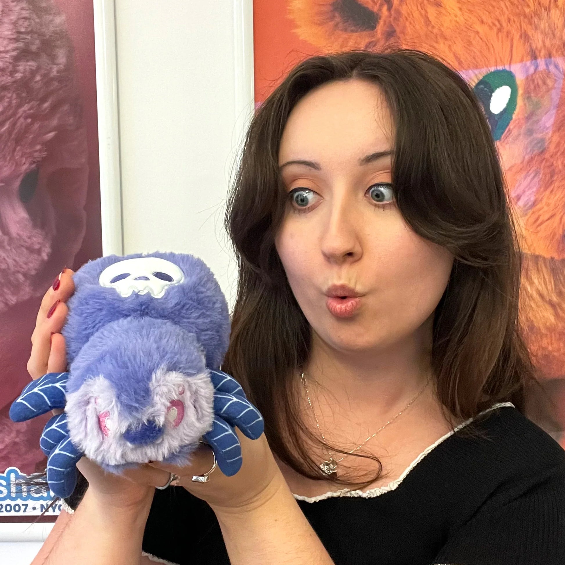 A woman holding a blue and purple plush spider toy with a white skull face, posing in front of a colorful background.