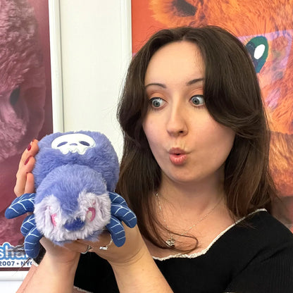 A woman holding a blue and purple plush spider toy with a white skull face, posing in front of a colorful background.