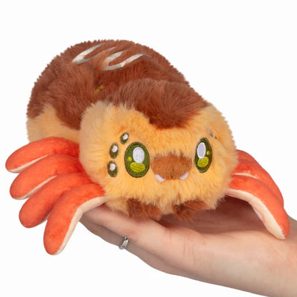 Squishable Alter Egos Series 20 Takoyaki Spider plush held in a hand, featuring golden-brown fur with white drizzle embroidery, orange tentacle legs, and green eyes.