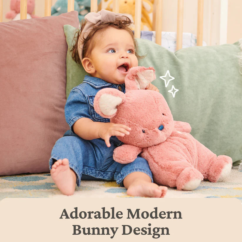 Baby holding a pink bunny plush toy in a crib with colorful pillows.