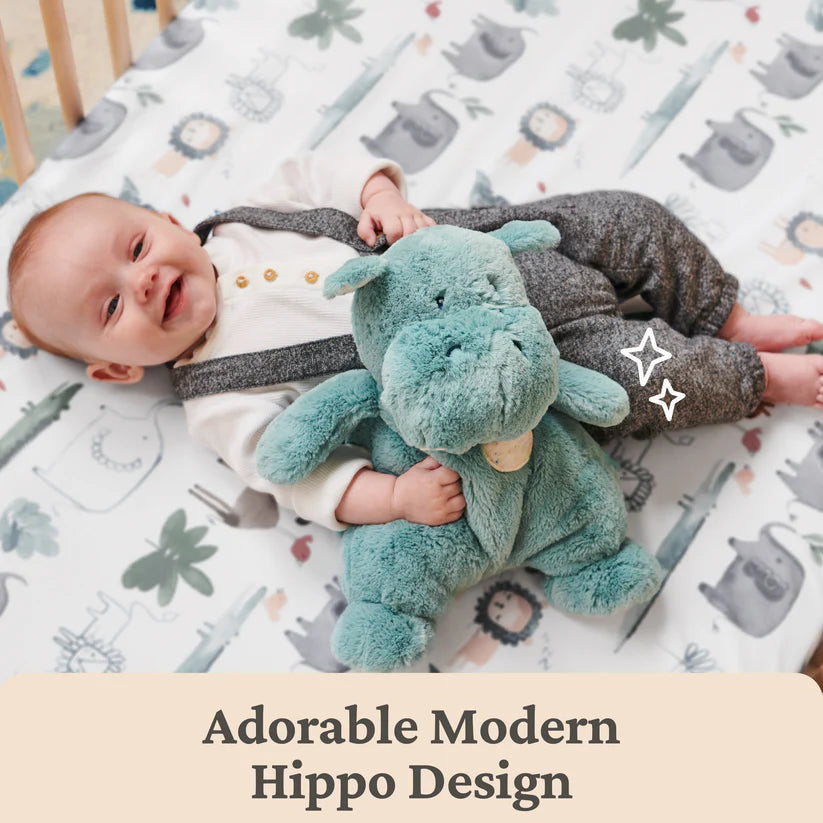 Baby lying on a crib with a plush hippo toy, surrounded by a patterned sheet.