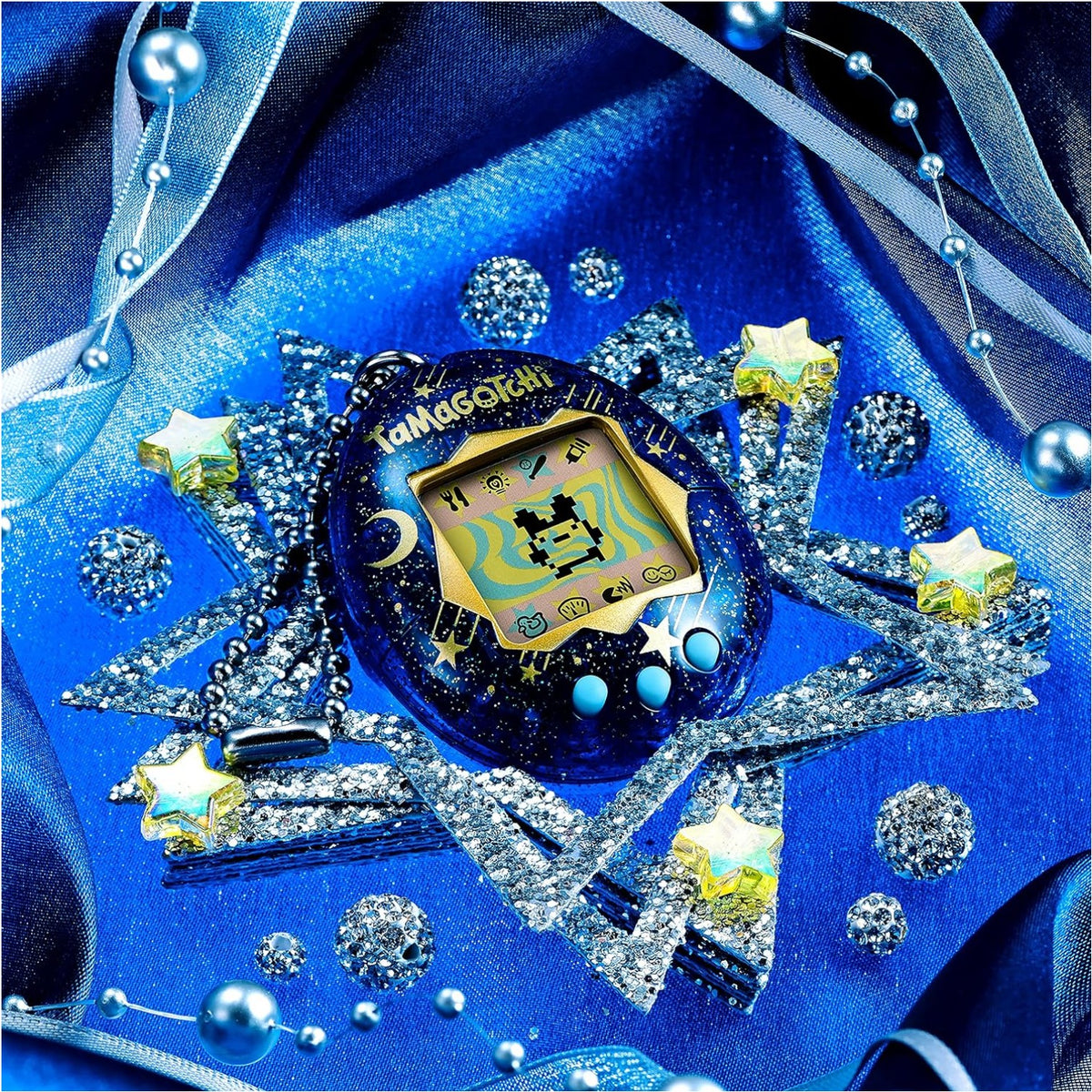 A blue Tamagotchi toy with a glittery space theme, surrounded by silver star-shaped accessories, iridescent stars, and blue beads on a shiny royal blue fabric background.