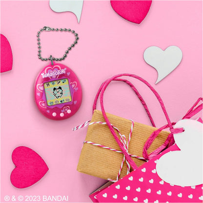 A pink Tamagotchi keychain toy with a silver ball chain, displayed on a pink background with heart-shaped cutouts, a wrapped gift box, and a pink heart-patterned bag. The Bandai copyright watermark is visible in the bottom-left corner.