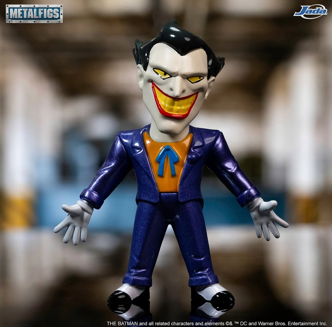 Metal-figs collectible figure of the Joker from The Batman, featuring a metallic purple suit, orange shirt, and white teeth, displayed against a blurred background.