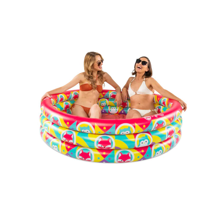 Two women sitting in an inflatable three-ring kiddie pool with a red bottom, featuring a colorful all-over print of Squishmallows characters on the sides.