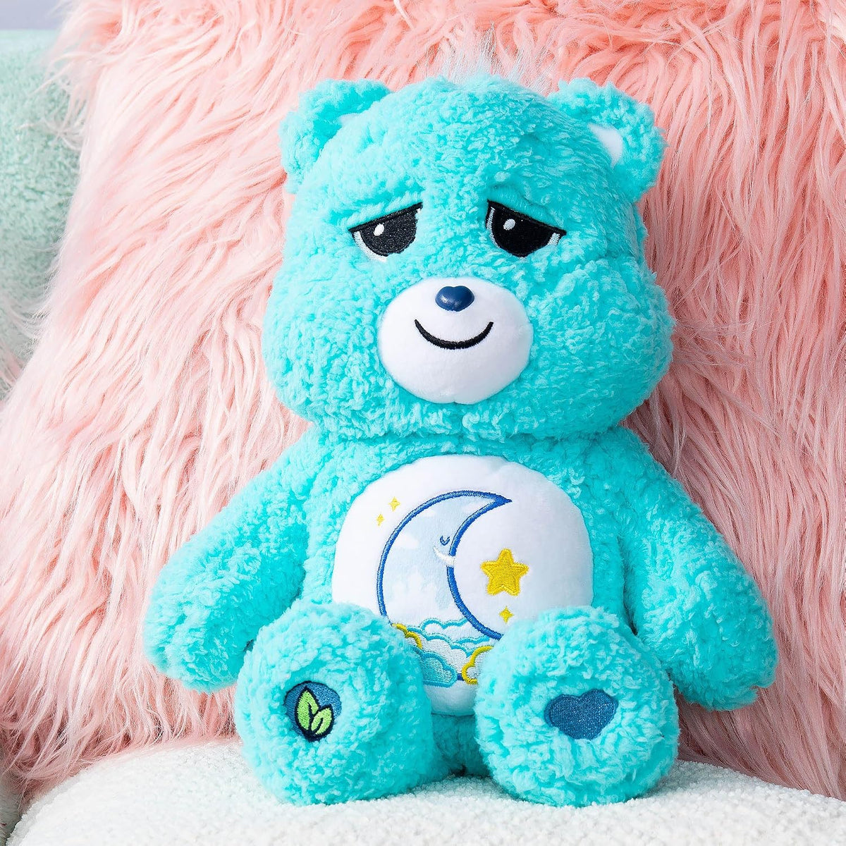Blue teddy bear with a white belly featuring a moon and star design, sitting against a pink fluffy background.