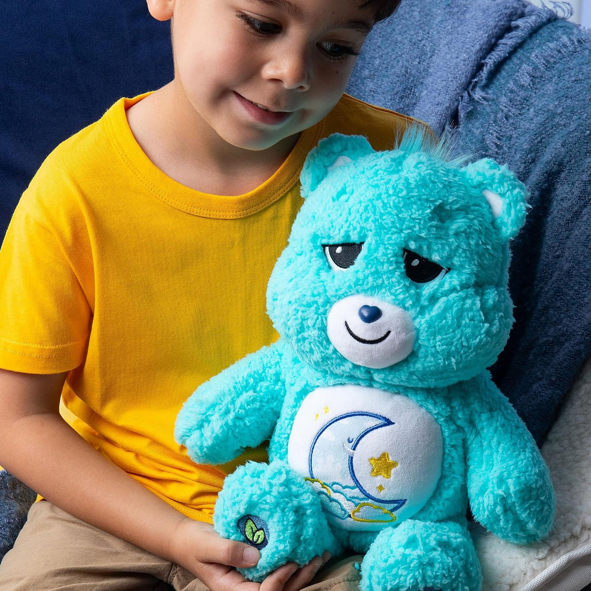 A young boy in a yellow t-shirt is smiling while holding a turquoise blue Care Bear plush toy with a crescent moon and star emblem on its belly, sitting on a blue couch.