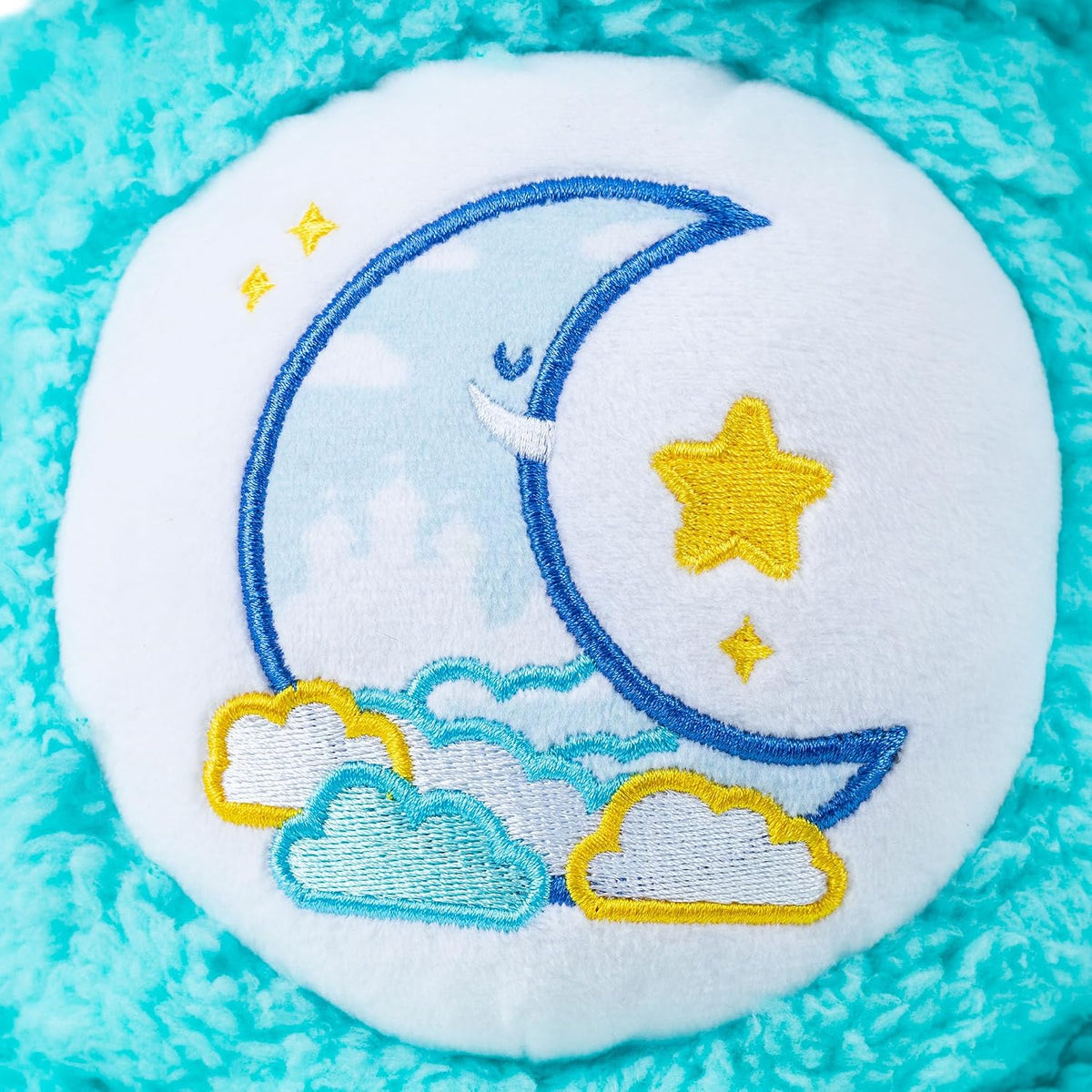 A close-up of a plush circular pillow featuring an embroidered crescent moon, yellow stars, and clouds in blue and yellow on a white background, surrounded by a turquoise textured fabric.