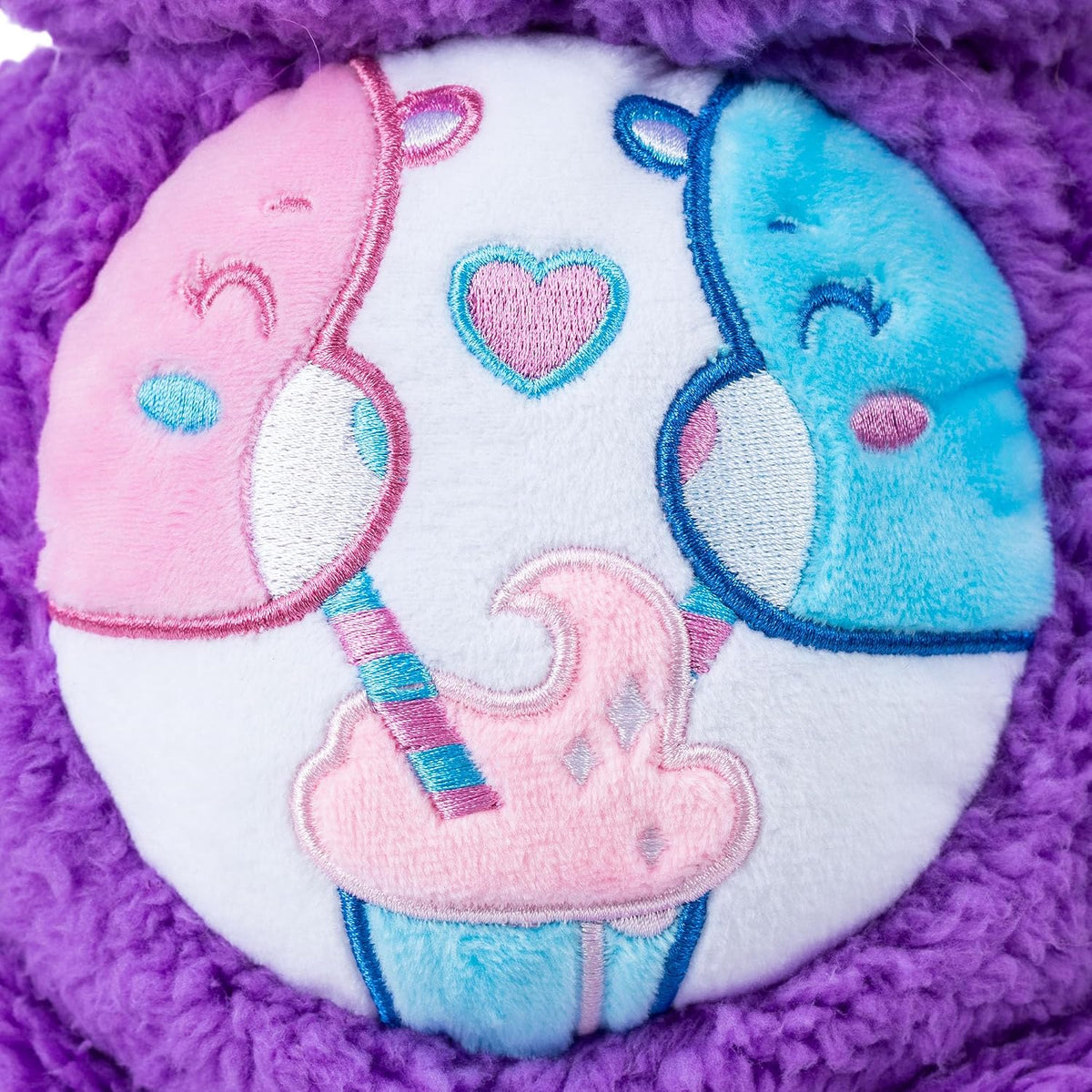 Close-up of a plush toy featuring a white circular patch with embroidered pastel characters: a pink bear on the left, a blue bear on the right, a pink heart in the center, and a pink-and-blue cupcake at the bottom, all surrounded by purple fuzzy fabric.
