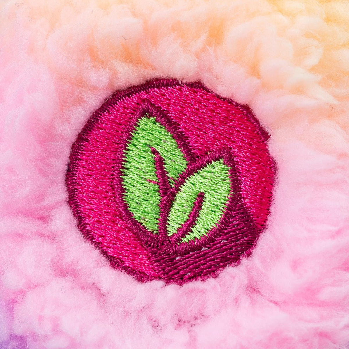 A close-up of a circular embroidered patch featuring two green leaves inside a bright pink circle, set against a soft, fuzzy pink and peach background.