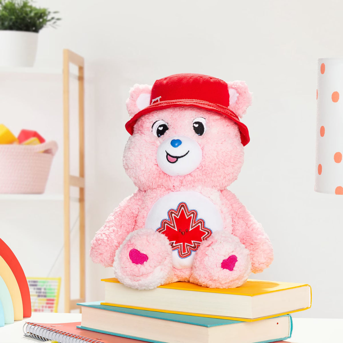 A pink plush teddy bear wearing a red cap and a white belly patch with a red maple leaf, sitting on stacked books in a bright indoor setting.