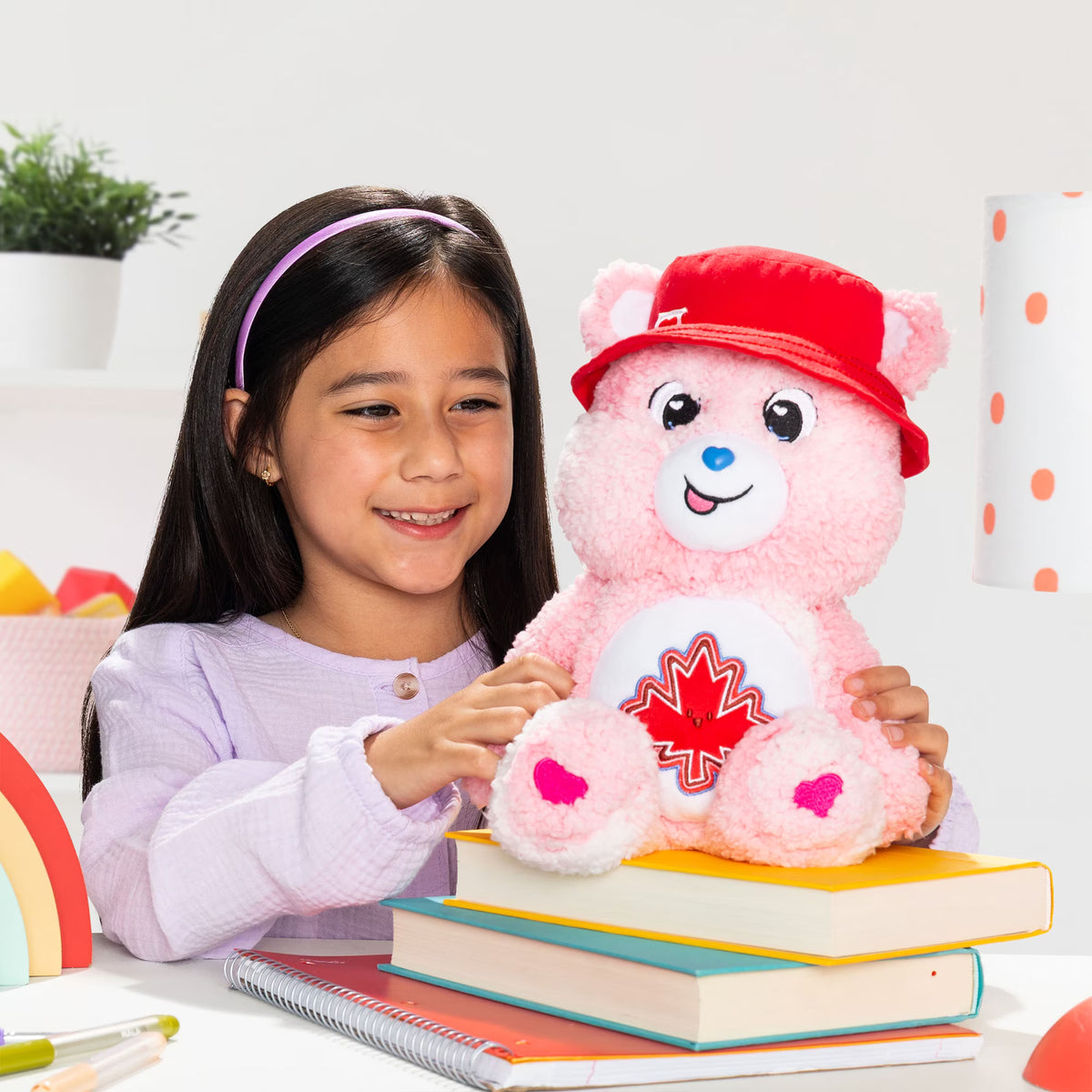 Young girl holding a pink teddy bear with a red cap and maple leaf design on a white background