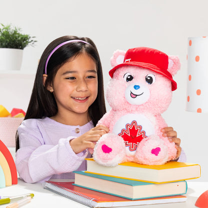 Young girl holding a pink teddy bear with a red cap and maple leaf design on a white background