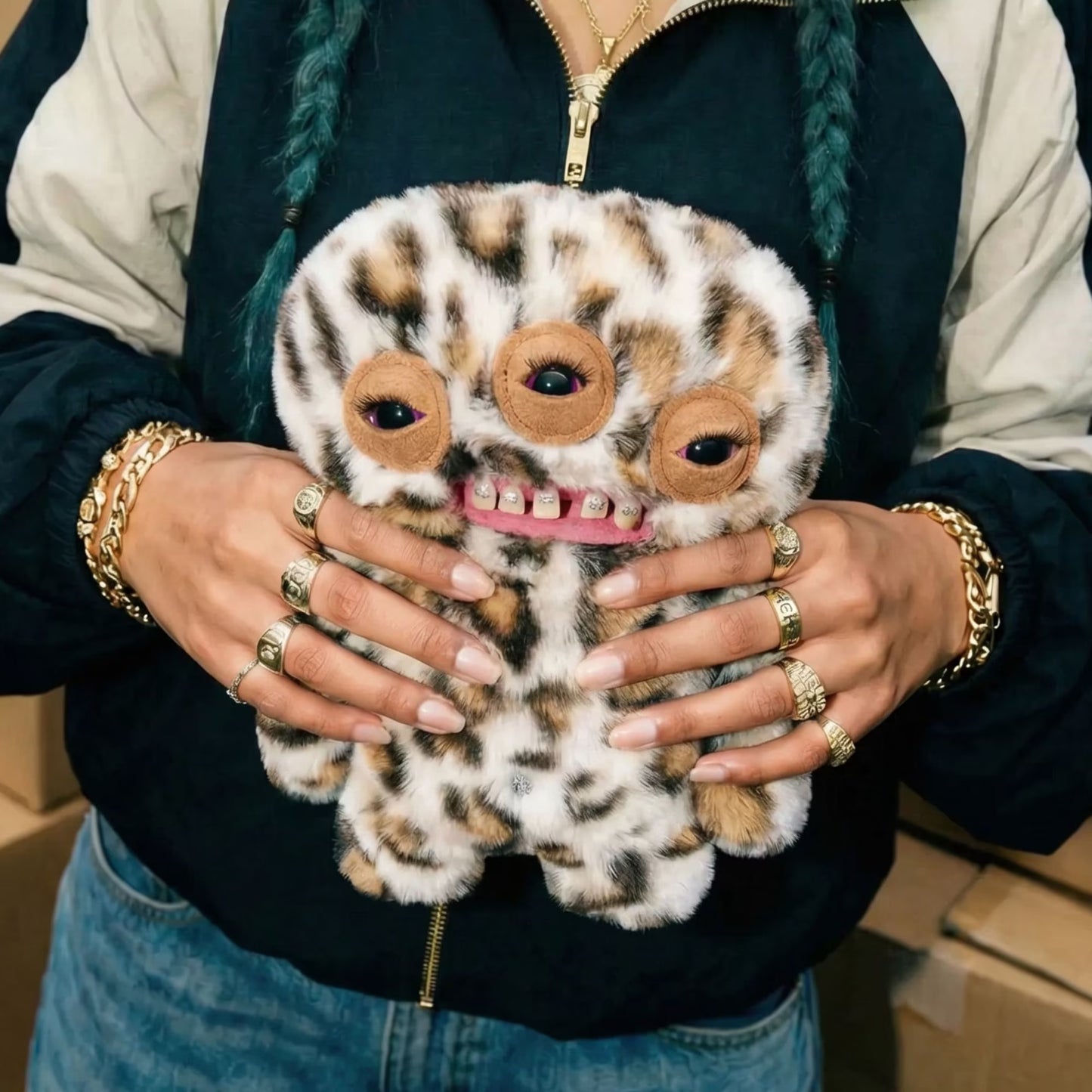 A person wearing a black and beige zip-up jacket holds a fuzzy monster plush toy with three large eyes, brown spots, and a pink mouth. The person is wearing multiple gold bracelets and rings.