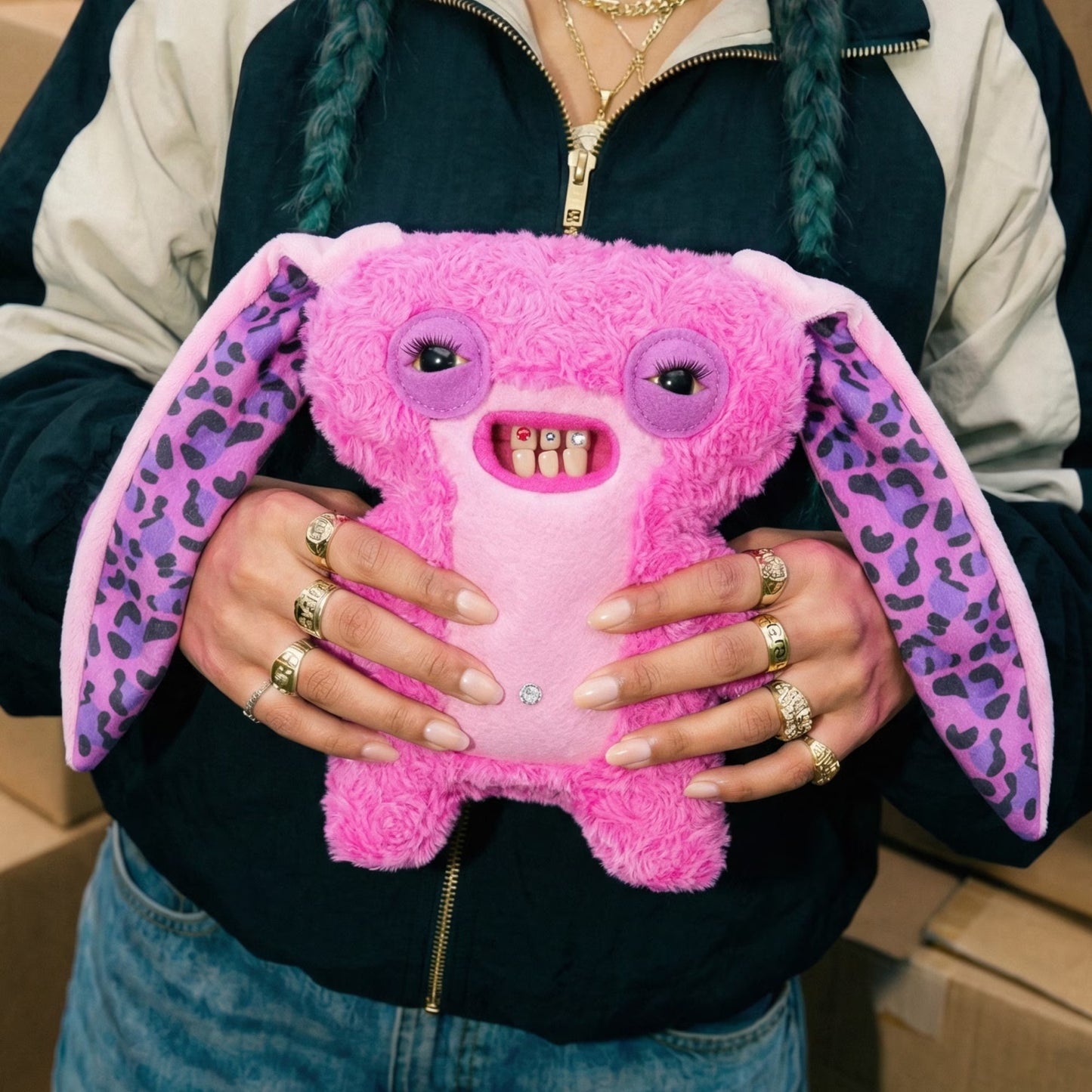 A person holding a pink plush toy with large floppy ears, purple leopard-print ears, and a toothy grin. The person is wearing a black and beige zip-up jacket and multiple gold rings.