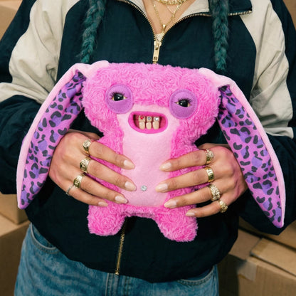 A person holding a pink plush toy with large floppy ears, purple leopard-print ears, and a toothy grin. The person is wearing a black and beige zip-up jacket and multiple gold rings.