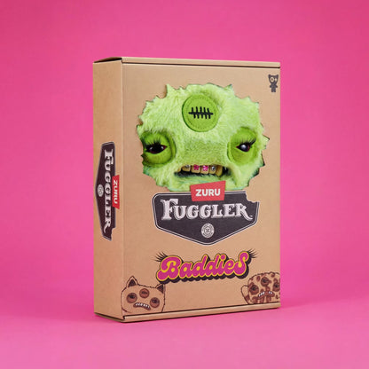 A cardboard product box for Zuru Fuggler Baddies, featuring a green stitched monster face cutout on the front, photographed against a solid pink background.