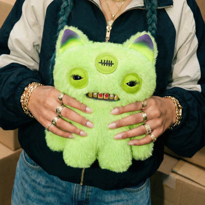 A person wearing a navy and white zip-up jacket holds a bright lime green fuzzy monster plush toy with purple inner ears, large eyes, and a stitched scar on the forehead.