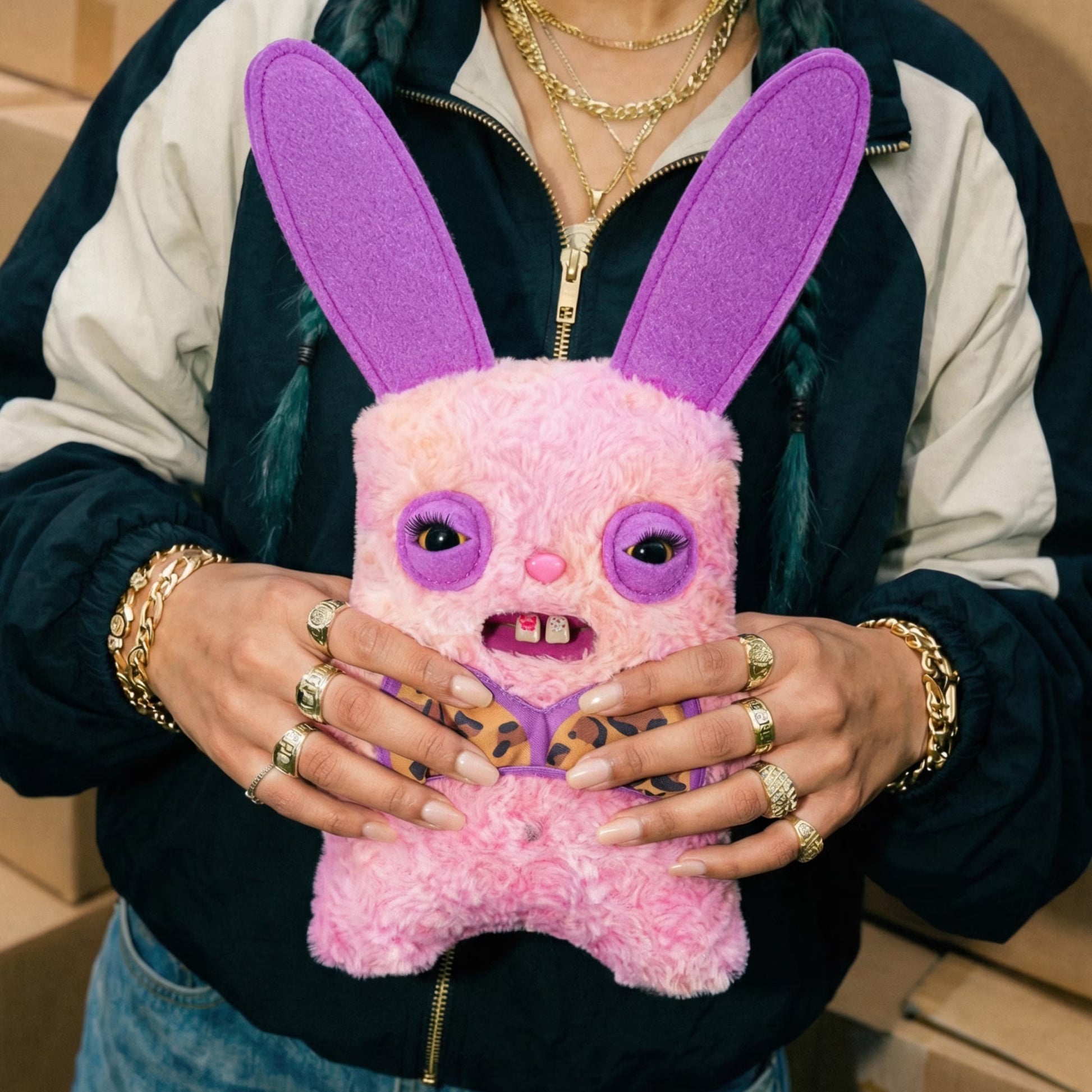 A person wearing a navy and cream zip-up jacket holds a pink fuzzy plush bunny toy with long purple ears, purple eye patches, and leopard-print details on the hands and chest.