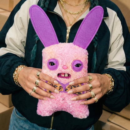 A person wearing a navy and cream zip-up jacket holds a pink fuzzy plush bunny toy with long purple ears, purple eye patches, and leopard-print details on the hands and chest.