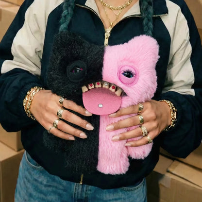 A person wearing a navy and beige zip-up jacket holds two fuzzy monster-shaped plush phone cases, one black and one pink, with one case open to show teeth and tongue details.