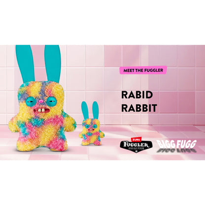 Marketing for A Fuggler Bigg Fugg 18-inch Rabid Rabbit plush toy, featuring rainbow tie-dye curly fur, large upright turquoise ears, squinting turquoise eyelids, and two large white teeth showing. Plush on a pink tile background with the words Bigg Fugg Rabid Rabbit and Meet the Fuggler.