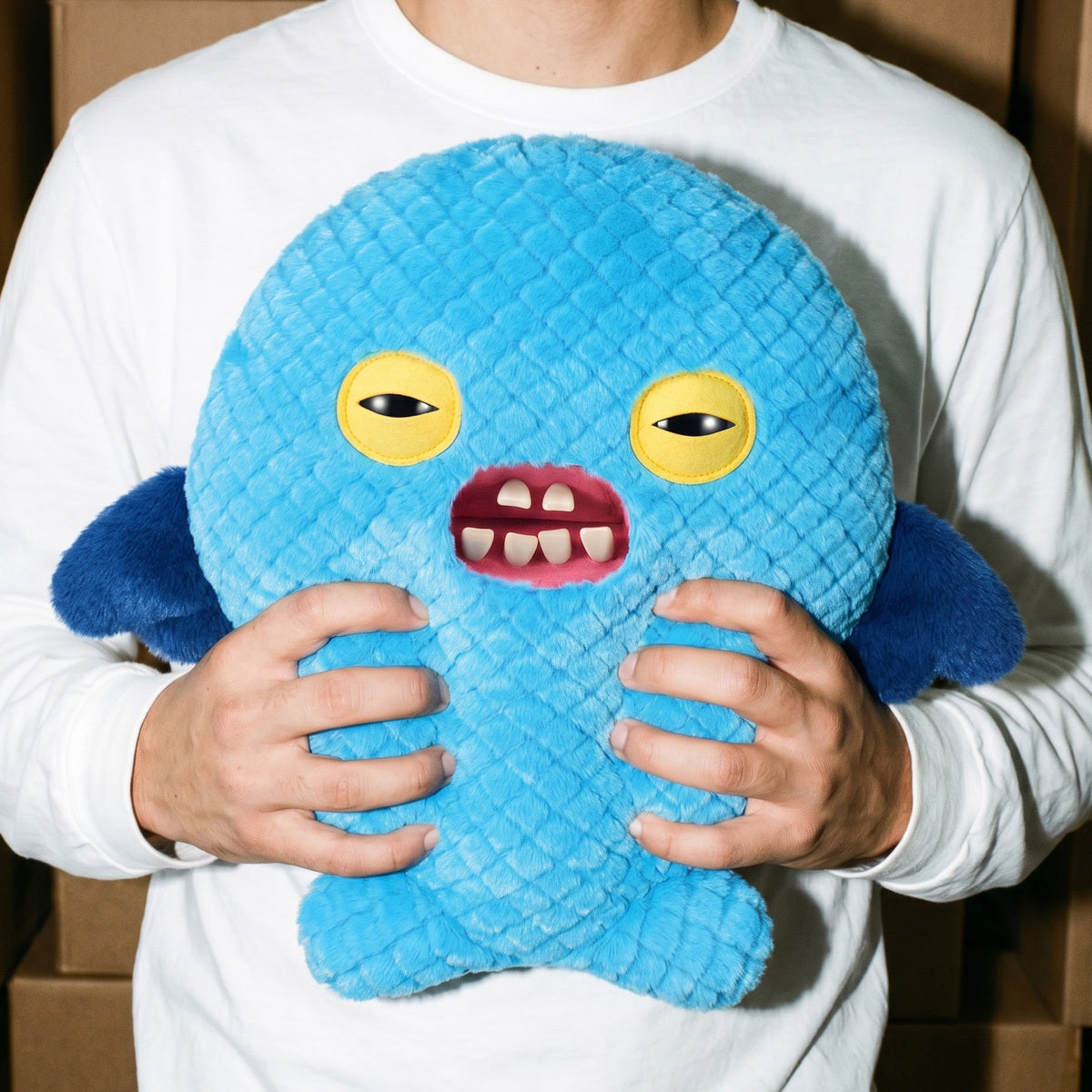 Person holding a Fuggler Bigg Fugg Cod Father 18-Inch Plush featuring a round blue textured body, yellow eyes, and unsettling human-like teeth.