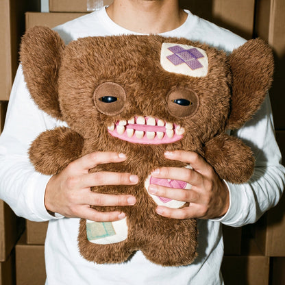 Person holding a Fuggler Bigg Fugg Grumpy Grumps 18-Inch Plush featuring fuzzy brown fur, human-like teeth, and large heavy-lidded eyes.