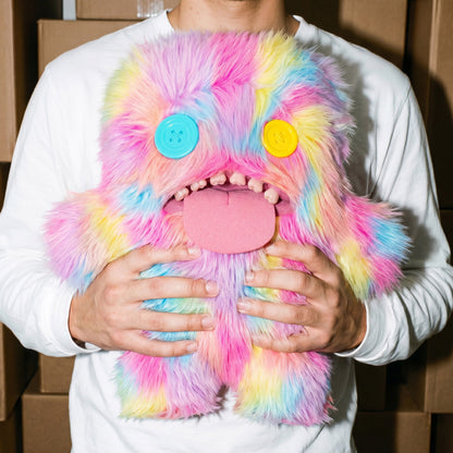 Person holding a Fuggler Bigg Fugg Oogah Boogah 18-Inch Plush featuring pastel tie-dye fur, mismatched button eyes, a pink tongue, and human-like teeth.