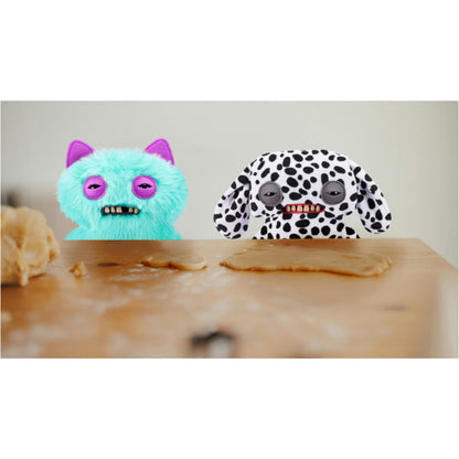 Two plush toys, one turquoise with pink ears and the other black and white spotted, sitting at a wooden table.