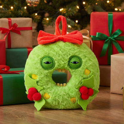 A green fuzzy Christmas ornament shaped like a wreath with a red bow, green eyes, and holly accents, displayed on a wooden surface with wrapped gifts and a Christmas tree in the background.