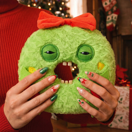 A person in a red knit sweater holds a Fuggler Festive Fuggs Series 2 Keith Wreathington 9-inch green wreath plush with human-like teeth and a red bow., holiday nail art, and a stitched open mouth showing small teeth.