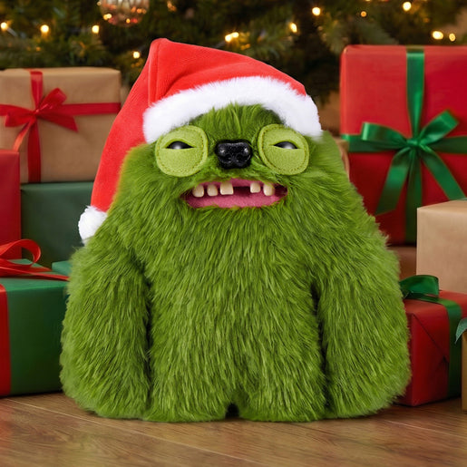 A Fuggler Festive Fuggs Series 2 green Sasquinch 9-inch plush showing wide human-like teeth and wearing a red Santa hat posed among wrapped holiday gift boxes on a wooden surface with a blurred Christmas tree in the background.