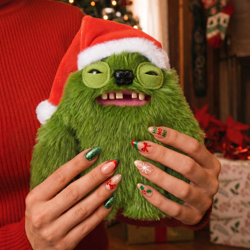 A Fuggler Festive Fuggs Series 2 green Sasquinch 9-inch plush showing wide human-like teeth and wearing a red Santa hat., held in both hands against a cozy holiday background with Christmas decorations and wrapped gifts.