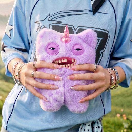 Person holding a Fuggler Fugglercorns Mr Screech plush toy with half solid and half spotted lavender fluff, a metallic pink unicorn horn, sleepy eyes, and a wide open mouth full of human-like teeth.