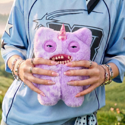 Person holding a Fuggler Fugglercorns Mr Screech plush toy with half solid and half spotted lavender fluff, a metallic pink unicorn horn, sleepy eyes, and a wide open mouth full of human-like teeth.