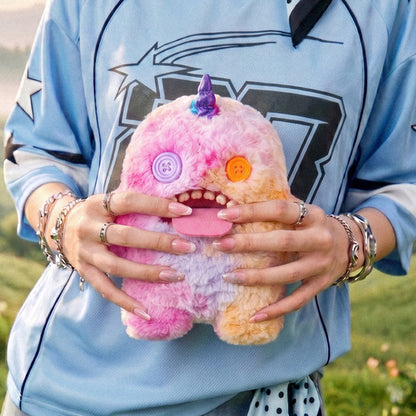 Person holding a Fuggler Fugglercorns Oogah Boogah plush toy with pastel tie-dye fur in pink, purple, and orange, a metallic purple unicorn horn, mismatched button eyes, a large pink tongue sticking out, and human-like teeth.