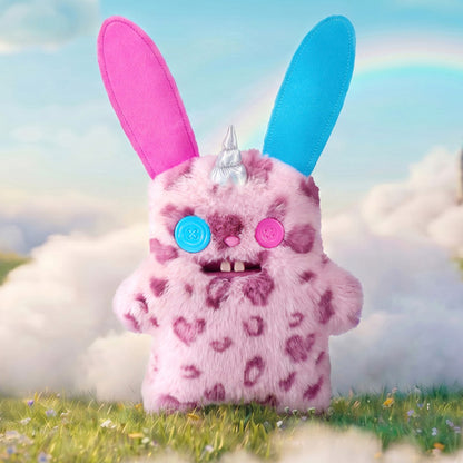 Fuggler Fugglercorns Rabid Rabbit plush toy with pink and dark purple spotted fur, tall ears with blue and pink insides, a silver unicorn horn, mismatched button eyes, and two large buck teeth in a fantasy setting with rainbow and clouds